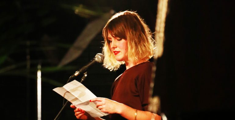 Young woman in long burgundy dress reading at a microphone
