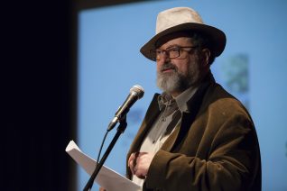 Middle aged man wearing a fedora reading at a microphone
